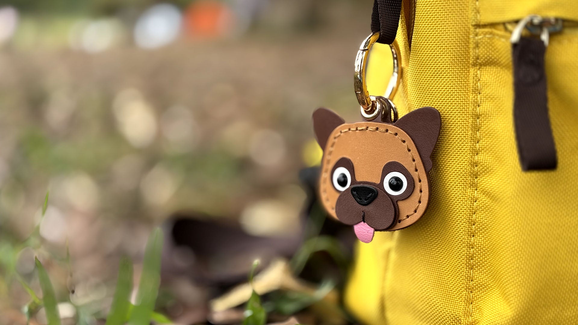 Handmade leather French Bulldog AirTag holder keychain, hanging on a yellow backpack outdoors