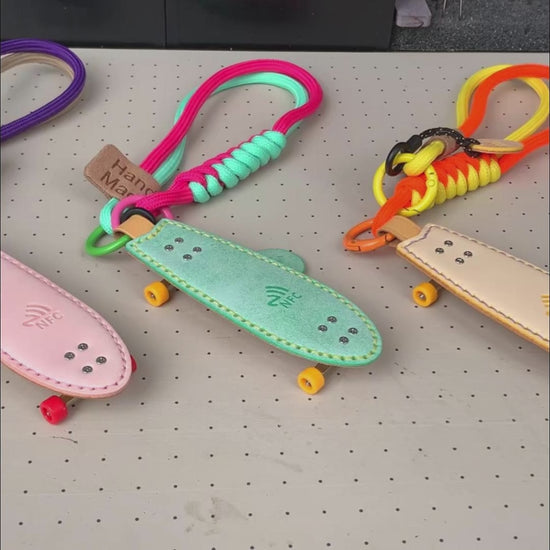 Video showcasing the three vibrant color options (Pink, Green, Yellow) of the handmade Waxy Veg-Tan leather Fishtail Skateboard Keychain, highlighting the built-in NFC logo, paracord straps, and mini fingerboard details.