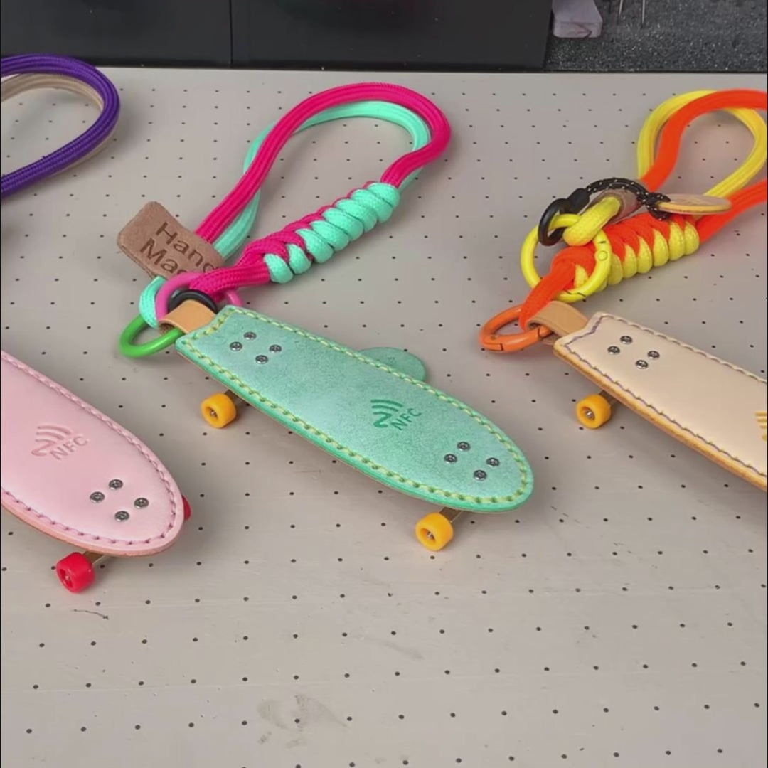 Video showcasing the three vibrant color options (Pink, Green, Yellow) of the handmade Waxy Veg-Tan leather Fishtail Skateboard Keychain, highlighting the built-in NFC logo, paracord straps, and mini fingerboard details.