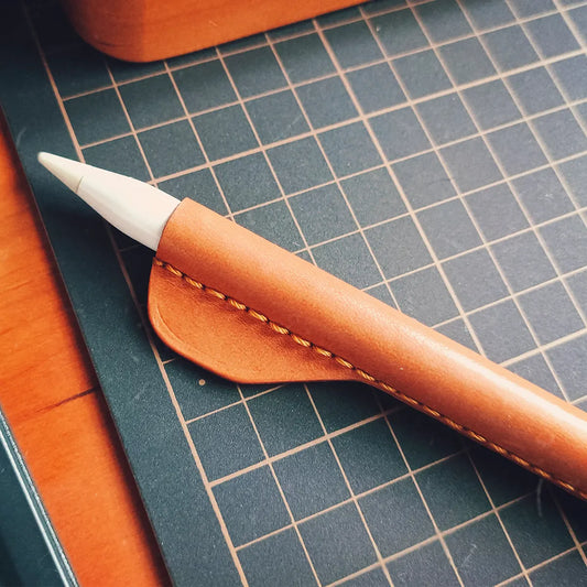 A premium saddle brown handcrafted leather Apple Pencil grip featuring a unique fish-fin ergonomic thumb rest on a studio work mat.